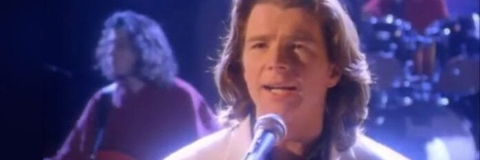 O cantor Rick Astley cantando de forma emotiva com um grande coral gospel ao fundo durante o início dos anos 90.