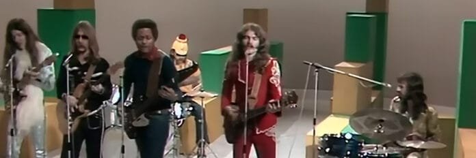 O vocalista Tom Johnston tocando guitarra e cantando com a banda The Doobie Brothers em um palco nos anos 70