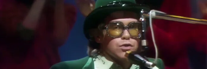 O cantor Elton John tocando piano usando seus tradicionais óculos extravagantes e roupas brilhantes na década de 70.